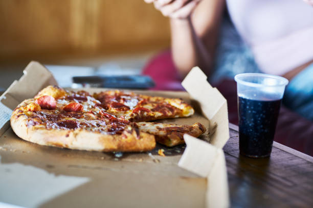close up of pizza in box with cola on coffee table while woman watches tv