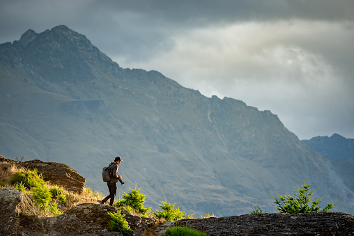 Young male photographer walking on Queenstown hill with mountain scenery in the background during golden hour sunset in Queenstown, South Island, New Zealand. Travel and photography concept