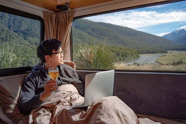 istock Young Asian man drinking beer and working with laptop computer on the bed in camper van with mountain scenic view through the window. digital nomad on road trip concept 1019214524