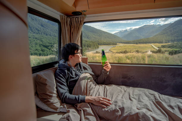 istock Young Asian man drinking beer and working with laptop computer on the bed in camper van with mountain scenic view through the window. digital nomad on road trip concept 1019214634