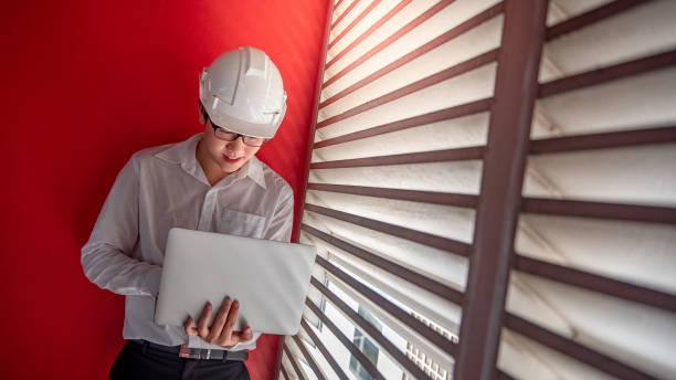 istock Young Asian male engineer or architect wearing protective helmet and glasses working with laptop computer with red wall in the background. Architecture, Engineering and building construction concepts 1019216798