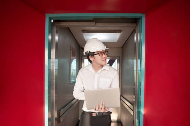 istock Young Asian male engineer or technician worker holding laptop computer in opened elevator door that surrounded by red wall. Mechanical engineering or building construction and maintenance concepts 1019216810