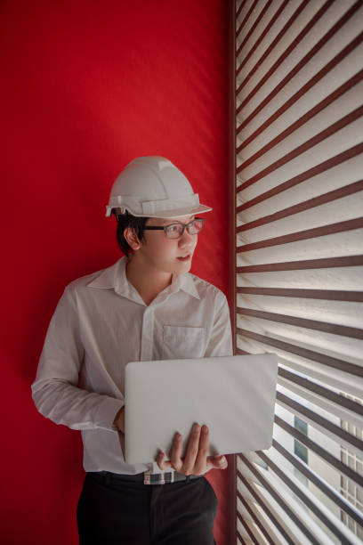 istock Young Asian male engineer or architect wearing protective helmet and glasses working with laptop computer with red wall in the background. Architecture, Engineering and building construction concepts 1019216812