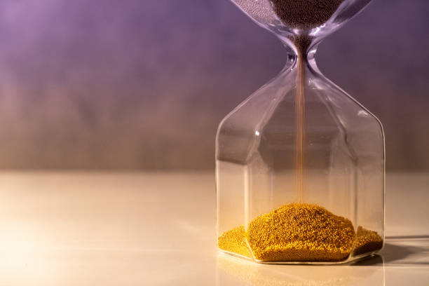 gold sand running through the shape of modern hourglass on white table.time passing and running out of time. urgency countdown timer for business deadline concept - hourglass time purple deadline imagens e fotografias de stock