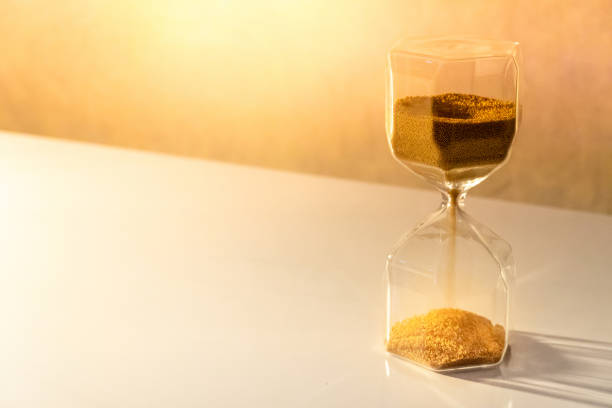 istock Gold sand running through the shape of modern hourglass on white table.Time passing and running out of time. Urgency countdown timer for business deadline concept 1019220030