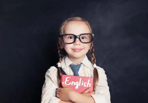 Cute child schoolgirl holding red book on chalkboard background. Speak english and learn language concept