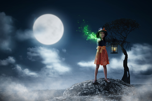 Pretty asian child girl in witch costume use magic wand with moonlight background