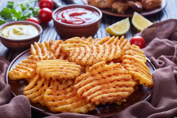 Crispy Potato Criss Cross Fries on a clay plates on a wooden table with mustard and tomato sauce dipping and sticky chicken wings at the background, view from above, close-up