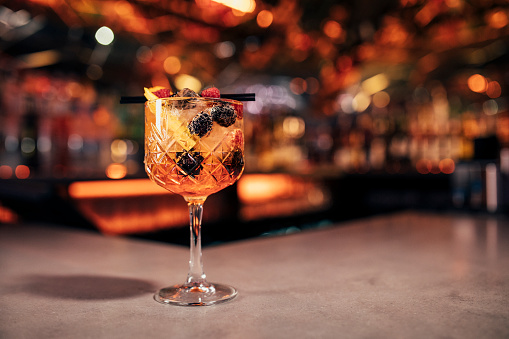 Front view of a Spritz cocktail on a bar counter. The background of the image is defocused lights and the back of the bar. This cocktail has blackberries and raspberries as a garnish on the cold drink.