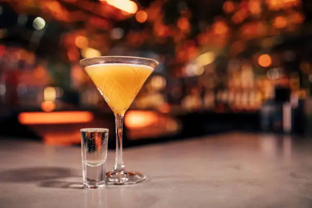 Front view of a Pornstar Martini cocktail with a shot of Prosecco  on a bar counter. The background of the image is defocused lights and the back of the bar. This cocktail has passionfruit as a garnish on the cold drink.