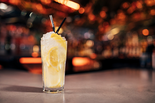 Front view of a Tom Collins cocktail on a bar counter. The background of the image is defocused lights and the back of the bar. This cocktail has a lemon slice as a garnish on the cold drink.