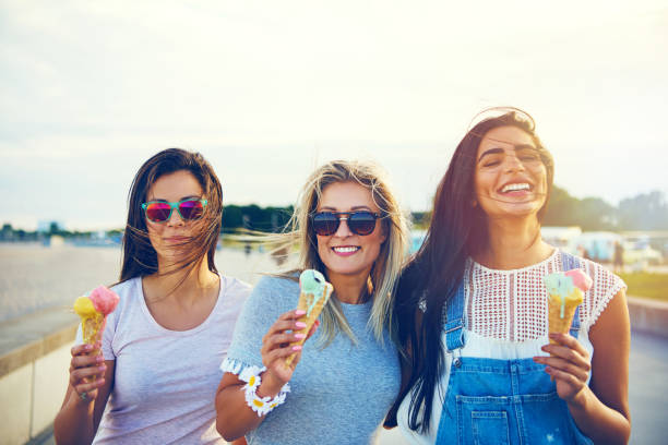 Three joyful young girlfriends on a promenade Three joyful young girlfriends on a promenade standing in a row smiling happily as they enjoy takeaway ice cream cones on summer vacation walking animation stock pictures, royalty-free photos & images