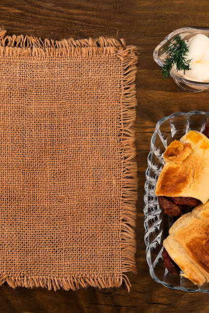 Fried meat sausages in a puff pastry bun, stacked in a glass plate, next to the gravy boat mayonnaise with a sprig of dill, linen napkin, all on a wooden background, top view.