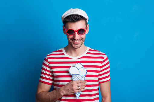 Portrait of a young man in a studio with sunglasses, a cap holding a paper ice cream on a blue background.