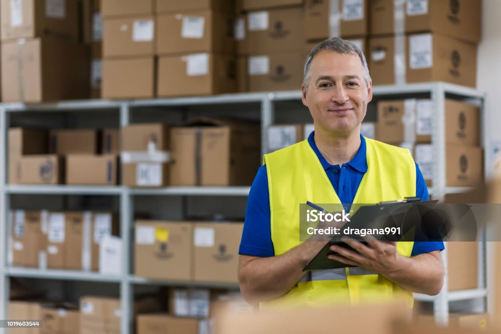 Warehouse worker holding clipboard in a large warehouse Warehouse worker holding clipboard in a large warehouse. Mature man looking up order details on a clipboard as he shops in a hardware warehouse for supplies , close up upper body Warehouse Worker Stock Photo