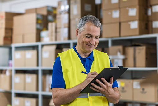 Man with clipboard in reflective safety vest at warehouse. Wholesale, logistic, people and export concept. Mature  man with clipboard smiling in warehouse