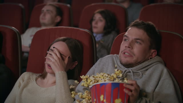 Young couple watching horror film in movie theatre. Guy sprinkle popcorn