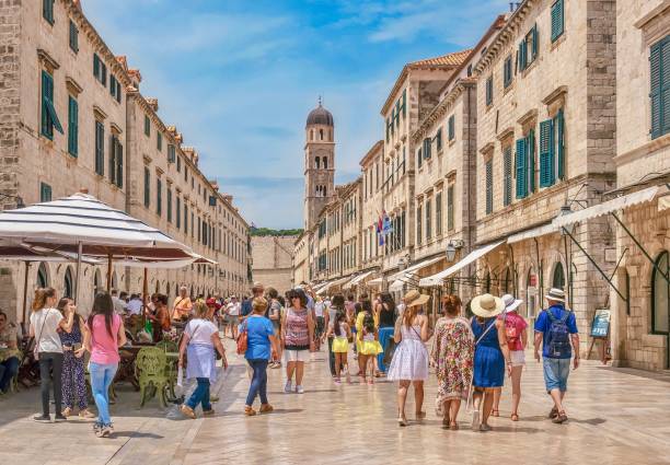 Tourists wearing summer clothing walk along the main street of old Dubrovnik, looking at the shops, restaurants and limestone buildings. stock photo