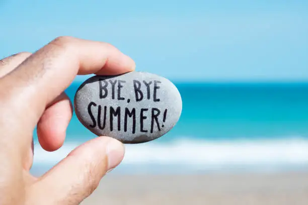 closeup of the hand of a young caucasian man on the beach, in front of the ocean, holding a stone with the text bye, bye summer written in it