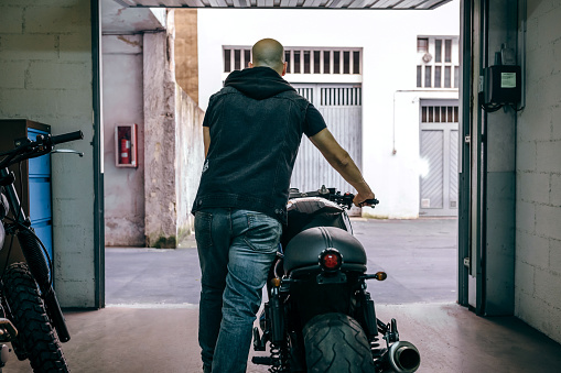Back view of man leaving the garage carrying a custom motorbike