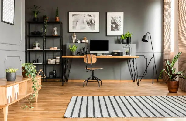 Striped rug near wooden cupboard in spacious grey workspace interior with posters above desk