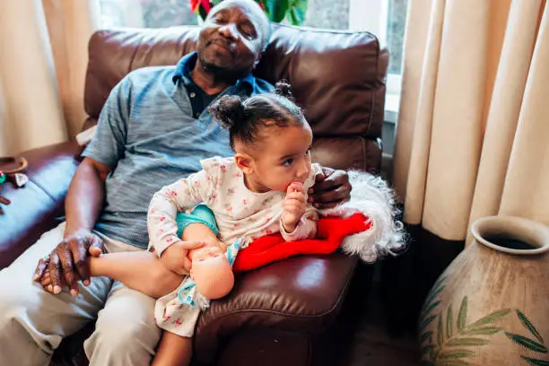 A little girl sits on the sofa with her grandfather at Christmas as he takes a nap.