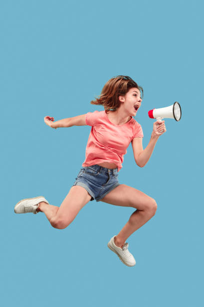 красивая молодая женщина прыгает с мегафоном изолированы на синем фоне - hip expressing positivity full blue стоковые фото и изображения