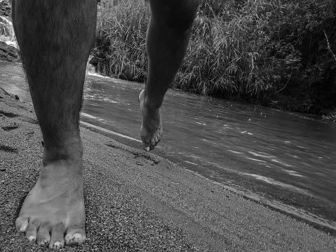 Black and white photograph of a man's legs walking on the sand, near a waterfall in a forest, in the rural city of Monte Belo, in Minas Gerais state, Brazil.