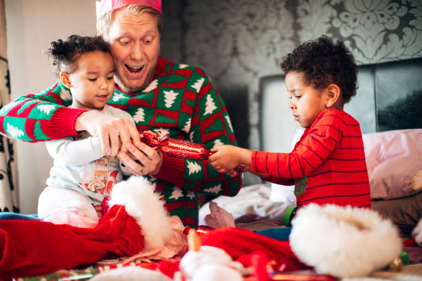 Siblings Pulling a Christmas Cracker with their Father at Christmas Time A brother and sister wearing pajamas excitedly pull a Christmas cracker whilst sitting on the bed with their father on Christmas morning. christmas cracker stock pictures, royalty-free photos & images