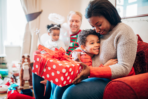 A little boy happily hugs his mother as she receives a gift from him on Christmas morning. His sister and father can be seen in the background.