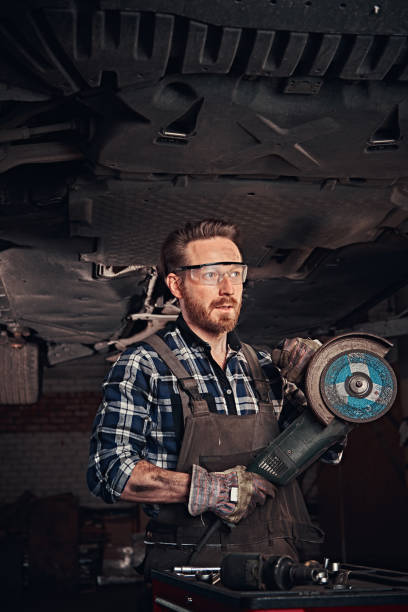 istock Mechanic in a uniform and safety glasses holds an angle grinder while standing under lifting car in a repair garage. 1019976754