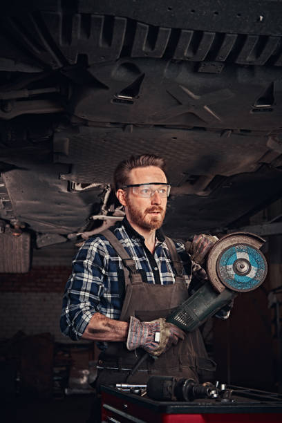 istock Mechanic in a uniform and safety glasses holds an angle grinder while standing under lifting car in a repair garage. 1019979684