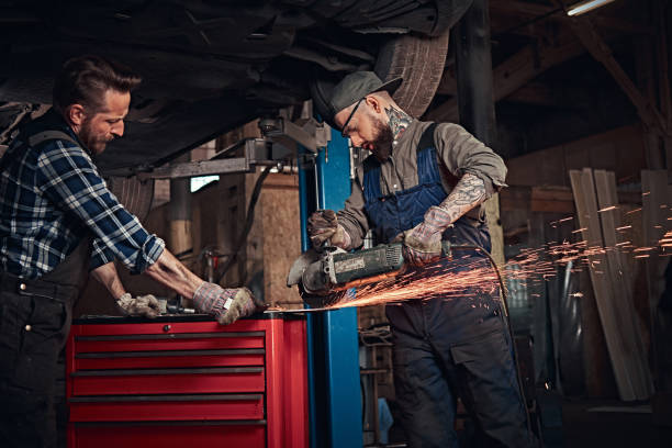 istock Two bearded auto mechanic in a uniform and safety glasses working with an angle grinder while standing under lifting car in repair garage. 1019981916