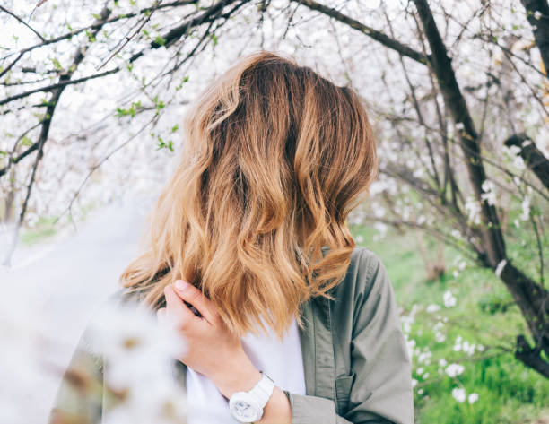 Young woman with fluttering blonde hair standing Young woman with fluttering blonde hair standing next to a blooming trees looking down. Candid lifestyle photo of fashionable female at spring day. wavy hair stock pictures, royalty-free photos & images