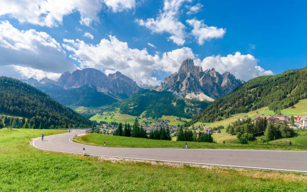 una vista de los alpes italianos y la ciudad de corvara (en primer plano hay un camino con dos ciclistas) - corvara fotografías e imágenes de stock