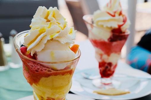 Close up of colorful ice cream sundaes. One in focus, one is blurry - in the background. Ice cream scoops with cream on it. Standing on white plates. Vivid background, summer mood. Side view.
