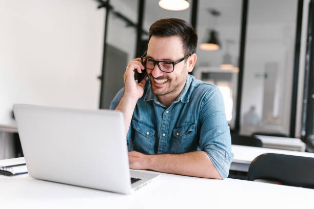 istock Smiling young man talking on smartphone while sitting in front of laptop. 1020064680