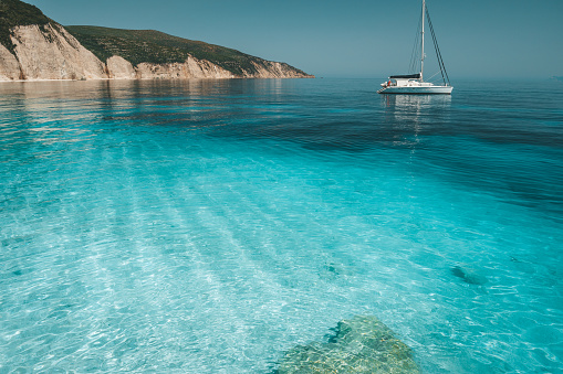 Azure blue lagoon with calm waves and drift sailing catamaran yacht boat. Rocky cliff coastline in background.