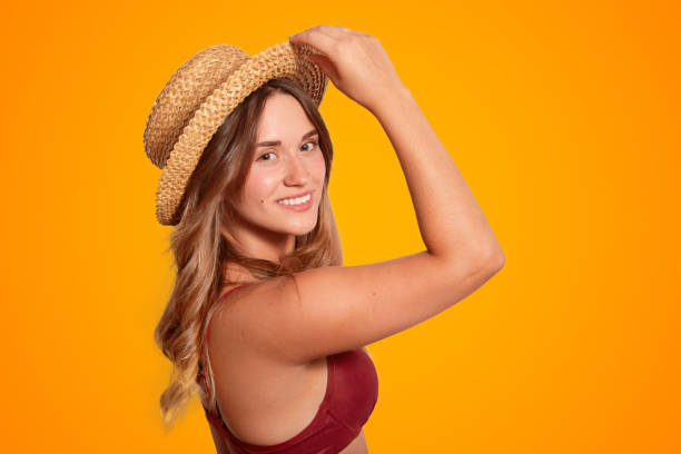 Sideways shot of pretty smiling young female model has pure healthy skin, wears summer hat and swimsuit, being in good mood after unforgettable rest in exotic country during summer vacation. tiro lateral de bastante sonriente joven modelo femenino tiene pura piel sana, se viste con sombrero de verano y traje de baño, estar en buen estado de ánimo después de un descanso inolvidable en país exótico durante vacaciones de verano.