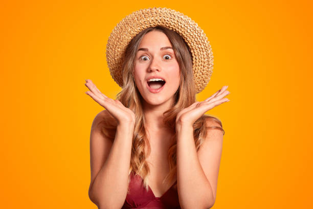 istock People, rest and emotions concept. Amazed surprised female in straw hat, keeps hands under chin, expresses surprisement, isolated over orange background. Woman tourist notices wonderful place 1020203420