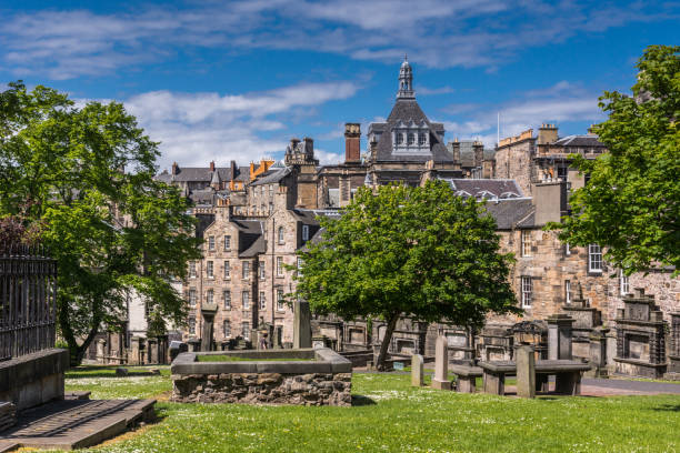 Kirkyard of Greyfriars Church, Edinburgh, Scotland, UK. Edinburgh, Scotland, UK - June 13, 2012: Graveyard of Greyfriars churk or kirk with green lawn, trees and tomb stones under blue sky. Backdrop is brown stone housing with tower of Central library. kirkyard stock pictures, royalty-free photos & images
