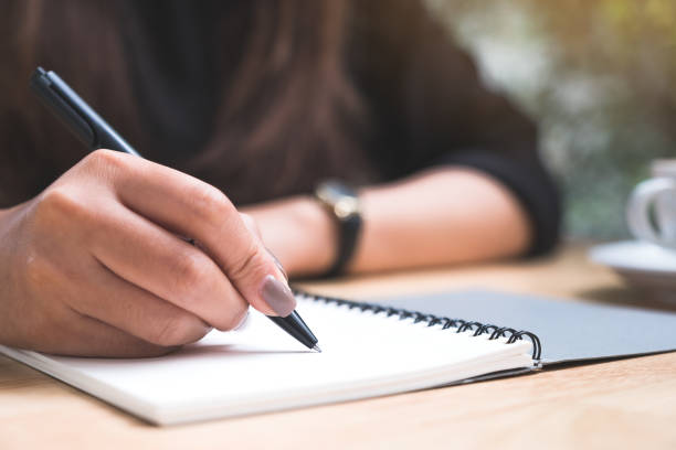 istock Closeup image of a woman's hand writing down on a white blank notebook with coffee cup on wooden table 1020325288