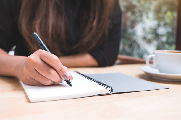 istock Closeup image of a woman's hand writing down on a white blank notebook with coffee cup on wooden table 1020325296