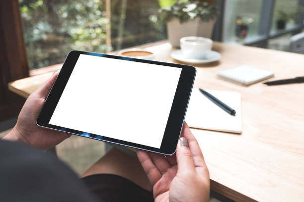 istock Mockup image of a woman's hands holding black tablet pc with white blank screen with notebook and coffee cup on table 1020325400