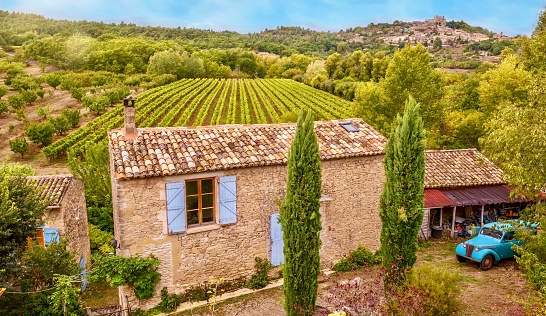 Rural Provence, France - October 13, 2016. Showing a traditional stone French farmhouse and grape vineyard. There is an old house with a blue shuttered window and clay tiled roof and a vintage blue car in the yard, with a green, rolling landscape in the background.