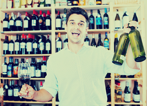 Cheerful man holding glass and bottle in winery section in store