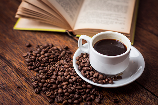 A cup of black coffee on a rustic wooden table can be seen next to a few coffee beans and a book.