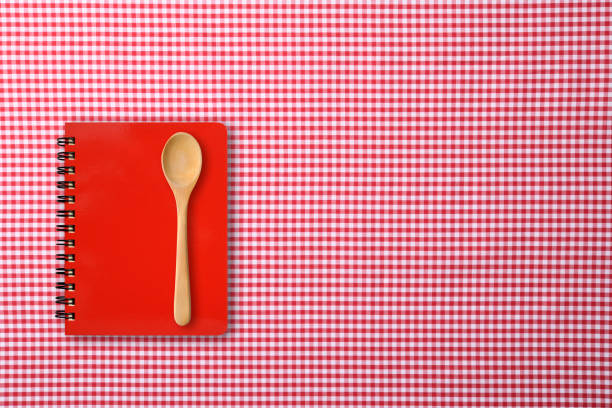 Overhead shot of closed red spiral notebook with wooden spoon on red gingham check tablecloth.