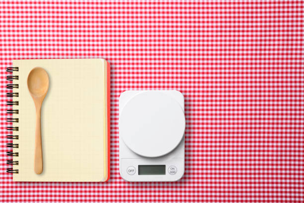 Overhead shot of kitchen digital food weight scale and spiral notebook  with wooden spoon on red gingham check tablecloth.