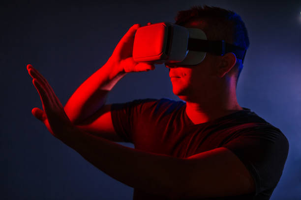 istock Amazed young smart casual man wearing virtual reality vr headset gesturing hands and looking at copyspace over black background dual color lighting 1020403524